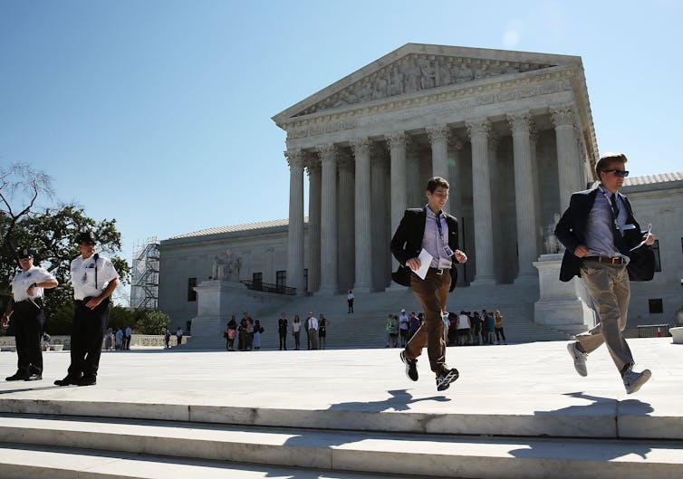 Two young men run in front of the Supreme Court building, with guards in white shirts looking on.