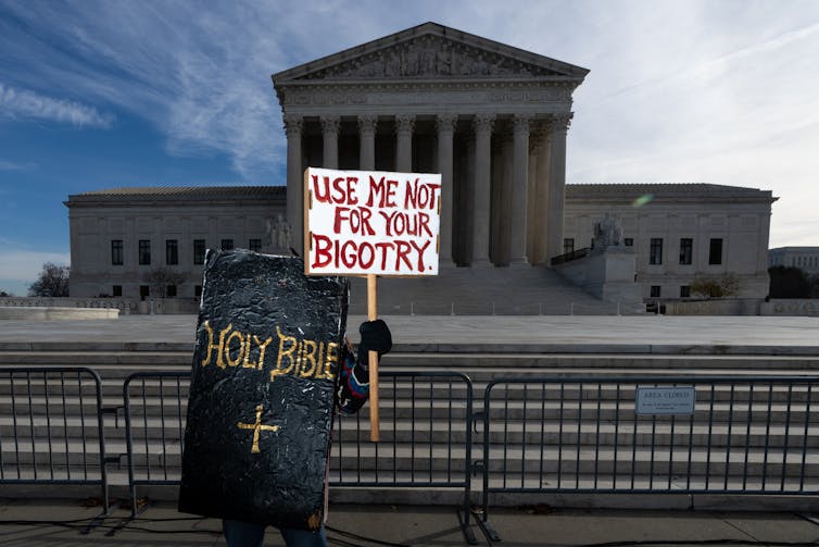 A person is blocked by a large fake holy bible which is black, and holds a sign that says 'use me not for your bigotry' in front of the Supreme Court