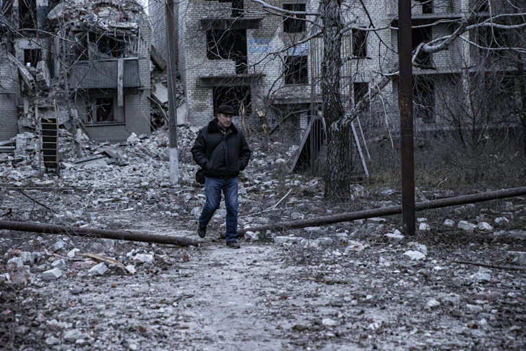 A man wearing a coat and hat is walking in the middle of concrete debris with his hands in his pockets.