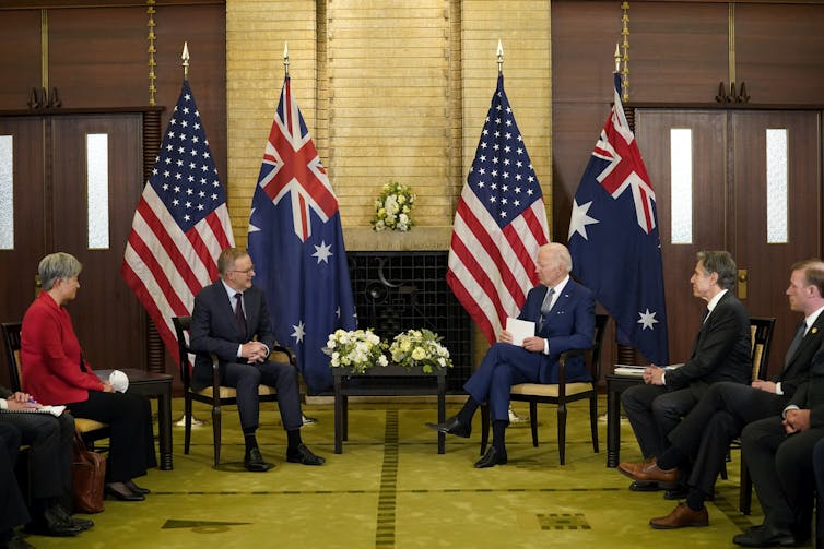 one woman and four men sit in front of flags