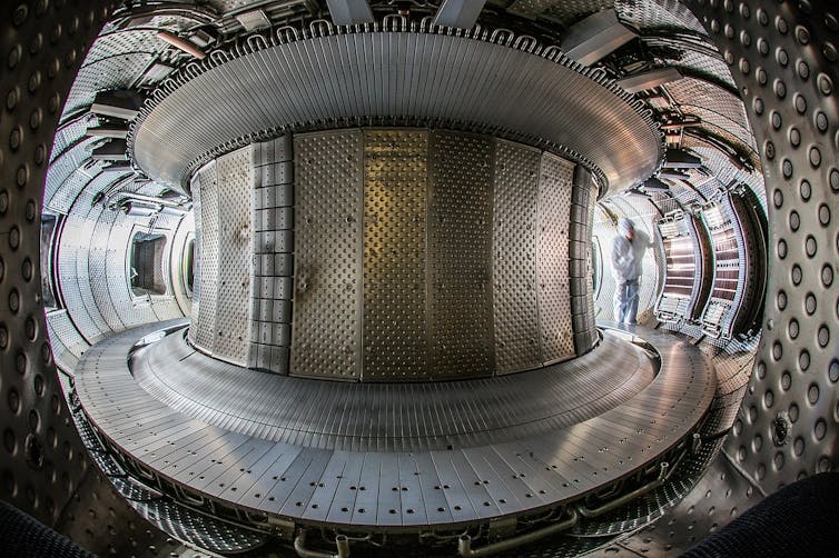 A fish-eye view of a metal-clad spherical room with a large column in the middle and a person in a white protective suit inspecting one of the walls