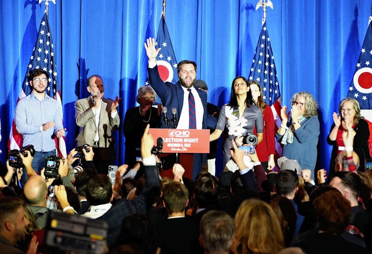 man waves at lectern surrounded by supporters