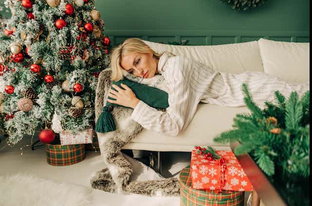 A woman lays on a couch next to her Christmas tree looking tired.