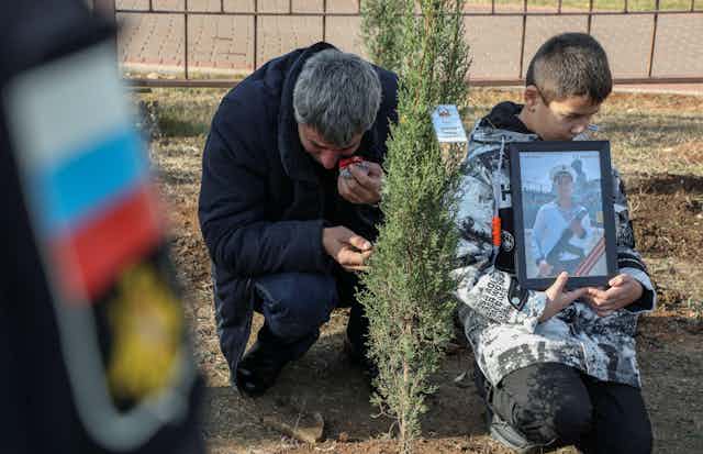 A Russian man cries as he plants a tree in memory of his son, Russian sailor Georgy Shakuro. A younbg boy holds a portrait of the dead sailor.nflict, mourns next to a boy holding Georgy's portrait during a tree planting ceremony in memory of fallen Russian service members in Sevastopol, Crimea, November 20, 2022. 