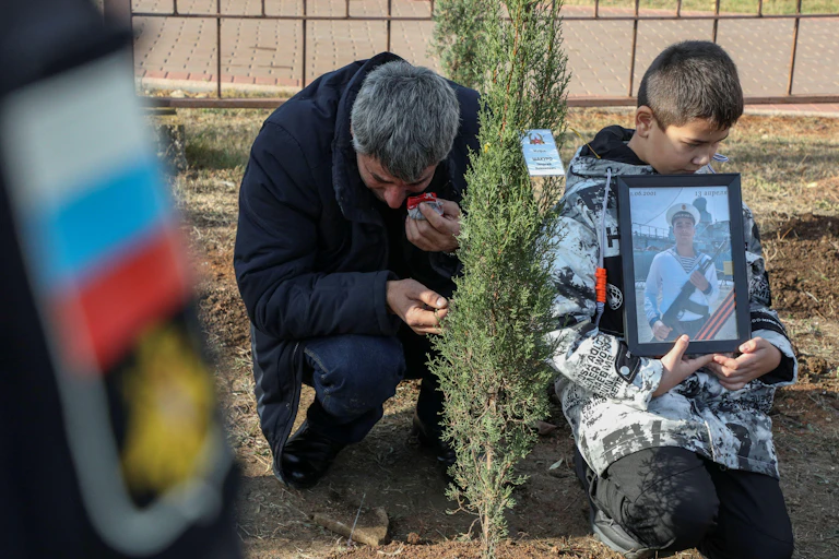 A Russian man cries as he plants a tree in memory of his son, Russian sailor Georgy Shakuro. A younbg boy holds a portrait of the dead sailor.nflict, mourns next to a boy holding Georgy's portrait during a tree planting ceremony in memory of fallen Russian service members in Sevastopol, Crimea, November 20, 2022.