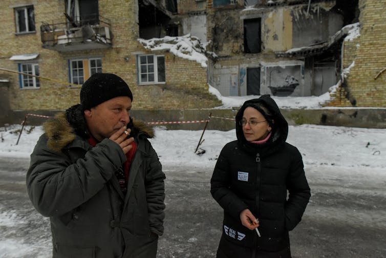 A man and woman smoking cigarettes outside a damaged apartment block covered in snow