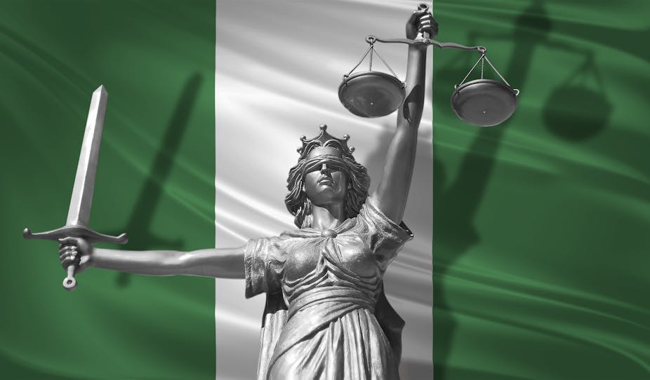 A statue in silver of lady justice, a woman with a blindfold on, holding up the scales of justice in one hand and a sword in the other, and standing in front of the flag of Nigeria. 