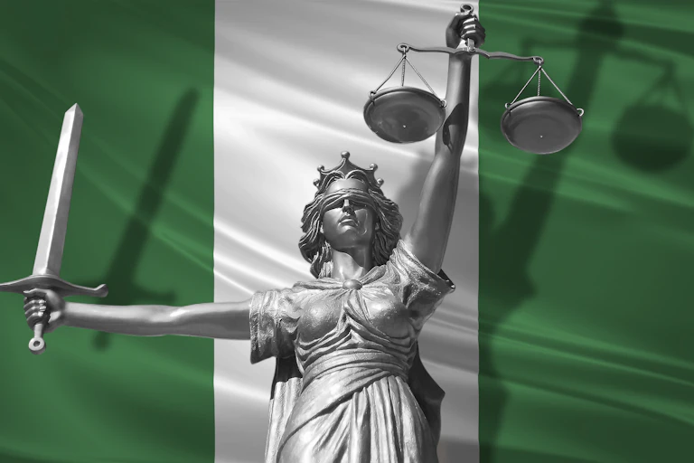 A statue in silver of lady justice, a woman with a blindfold on, holding up the scales of justice in one hand and a sword in the other, and standing in front of the flag of Nigeria.