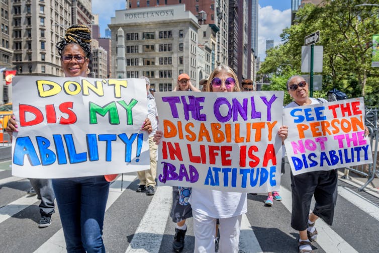 Three women stand next to each other in a city street, holding signs that say 'Don't dis my ability,' and 'the only disability in life is a bad attitude'