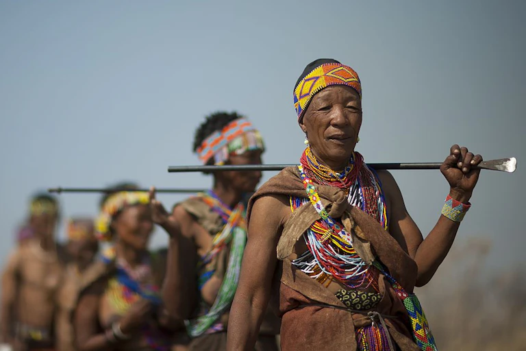 A group of people walk in single file, wearing traditional attire and carrying sticks.