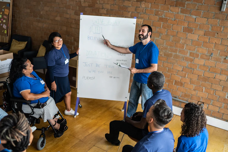 Man points to a whiteboard in a meeting where everyone in a diverse group wears blue shirts.