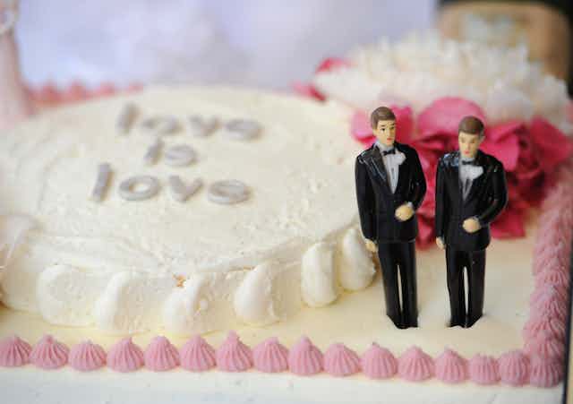 Two male figurines in formal suits stand on a white cake that says love is love