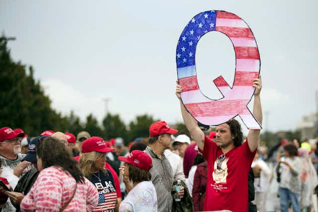 A dark-haired man in a red T-shirt holds up a Q sign.