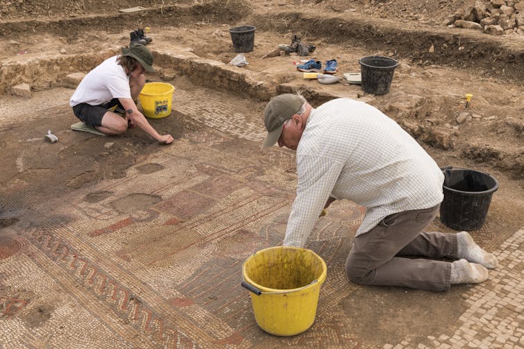 A man in the foreground and woman to the back of the shot bend down with buckets to excavate parts of the mosaic.
