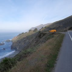 A yellow van parked on a steep road overlooking a large body of water