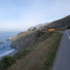 A yellow van parked on an escarpment road overlooking a large body of water