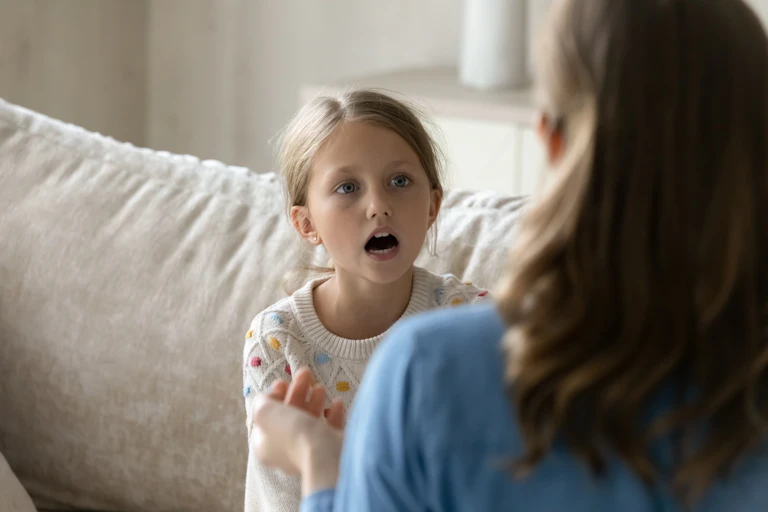 A young girl with blond hair speaks to an adult woman.