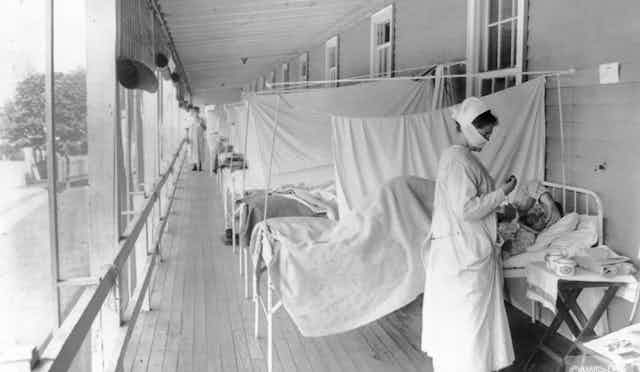 Patients lie on beds outdoors, separated only by sheets, while a nurse attends to a patient during the 1918 flu pandemic.