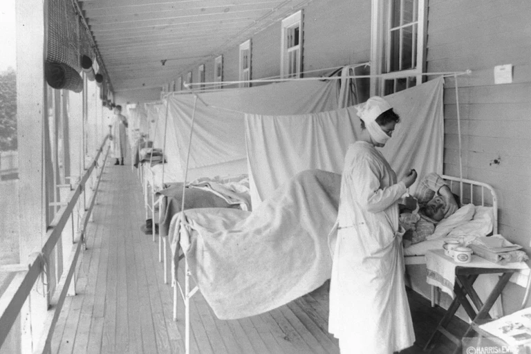 Patients lie on beds outdoors, separated only by sheets, while a nurse attends to a patient during the 1918 flu pandemic.
