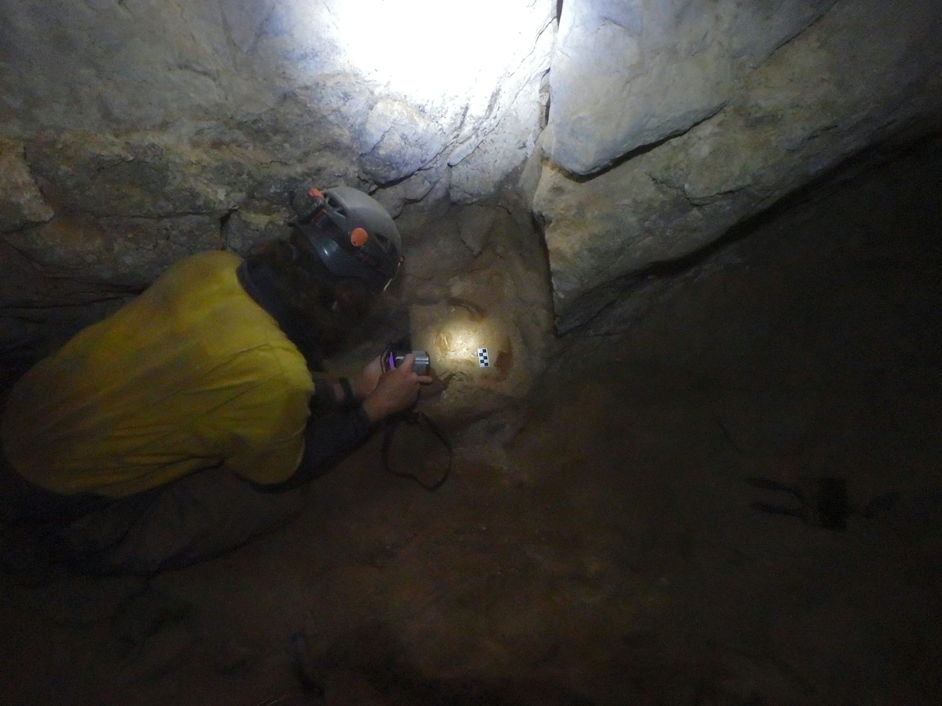 Dark photograph of a person in yellow shirt crouching next to rocks in a cave with a headlight on