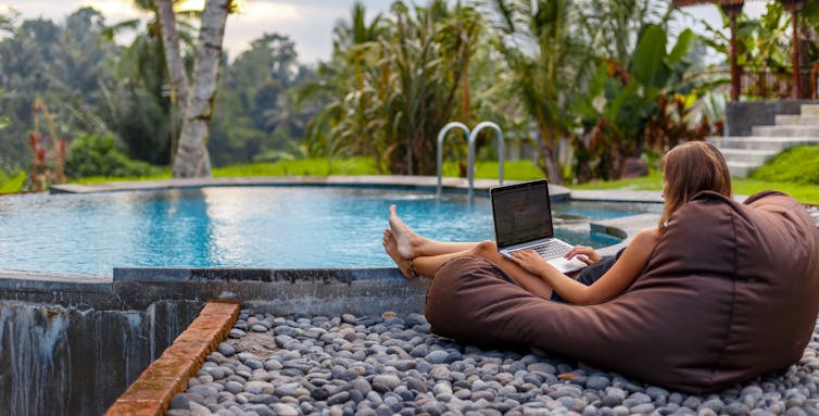 Woman with laptop sitting beside pool in tropical location.