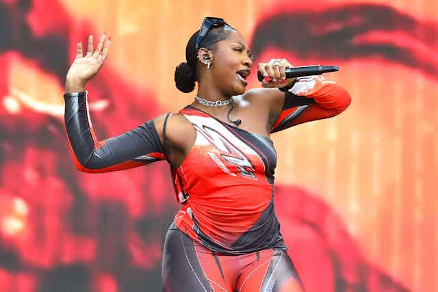 A glamorous woman on stage singing into a microphone. She has a body-hugging red, black and white outfit on and raises one hand in the air.