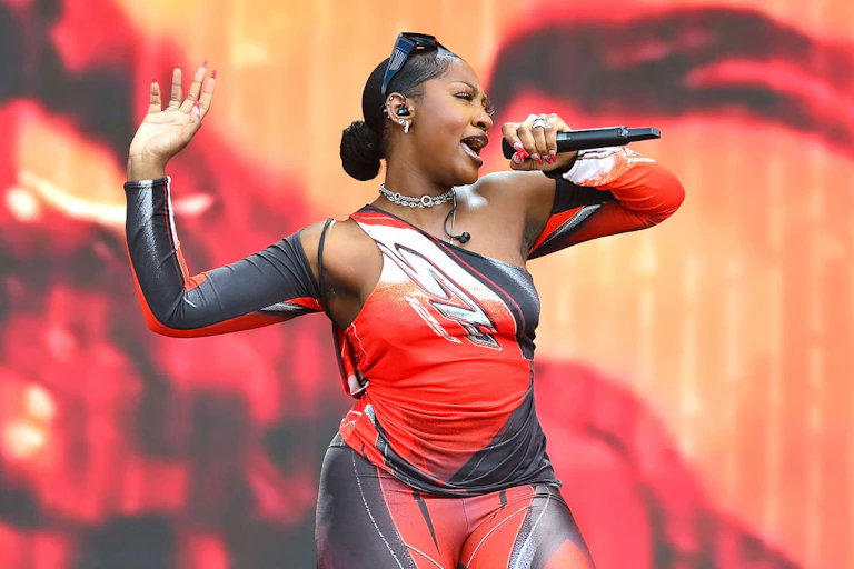 A glamorous woman on stage singing into a microphone. She has a body-hugging red, black and white outfit on and raises one hand in the air.