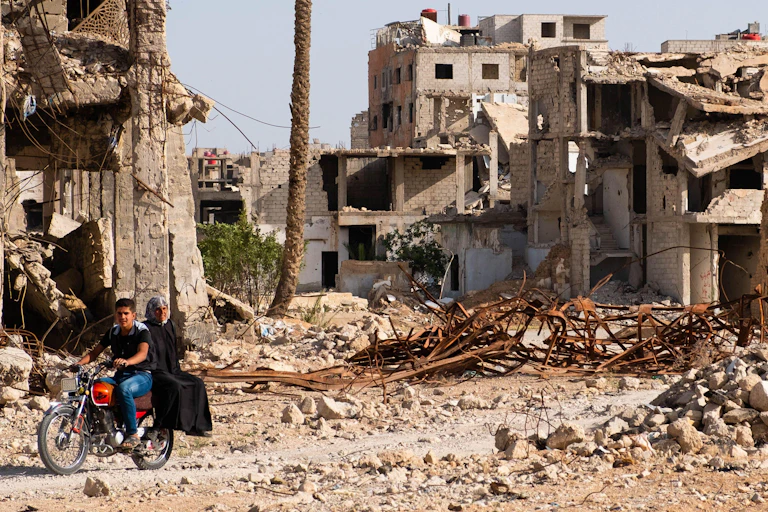 A man on a motorbike travels past ruined buildings.