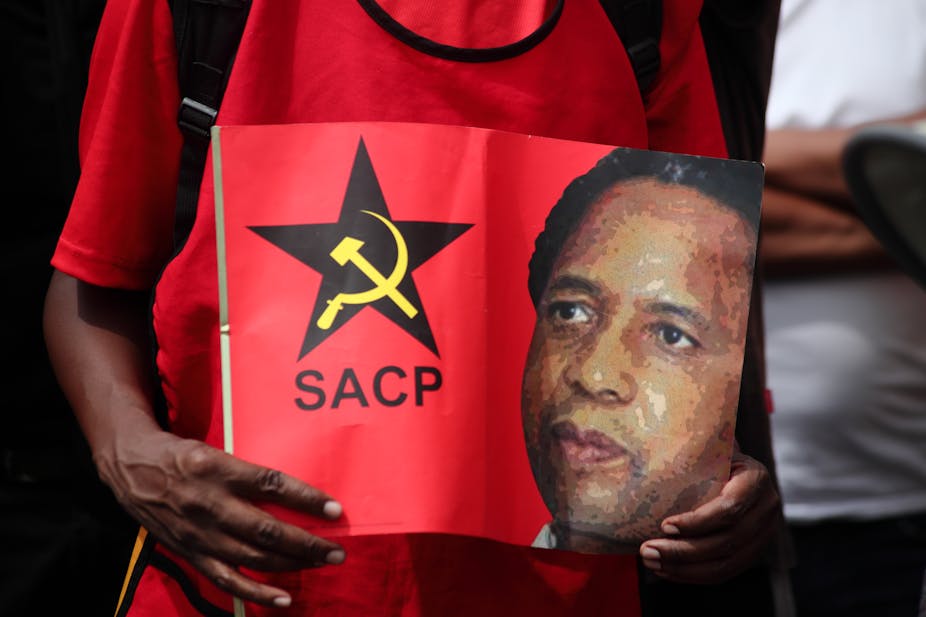 A man holds a placard showing the face of Chris Hani and the hammer and circle logo of the South African Communist Party.