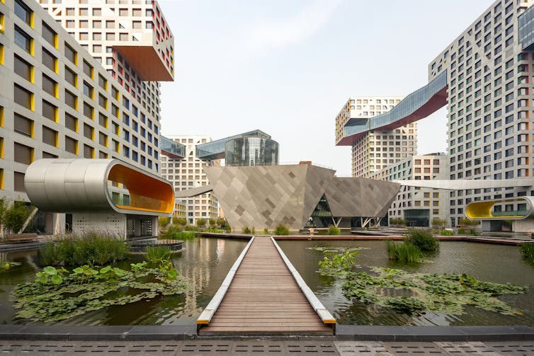 A wooden walkway seen extending through water and greenery leading towards a building complex.