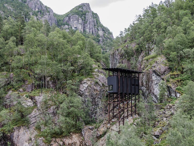 A thin structure appears to be on wooden stilts nestled against sheer rock.