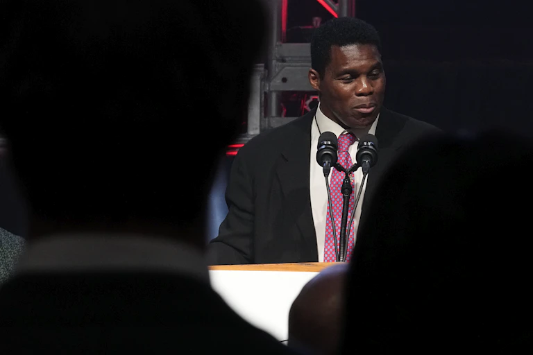 A man in a suit and pink tie talks into a microphone.