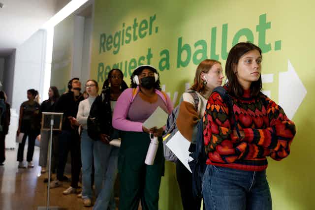 A row of young women stand next to a yellow wall with green writing that says 'Register. Request a ballot.'