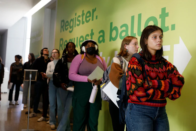 A row of young women stand next to a yellow wall with green writing that says 'Register. Request a ballot.'