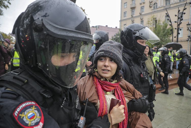 A young woman anti-war protester is detained by Russian security forces wearing riot gear.