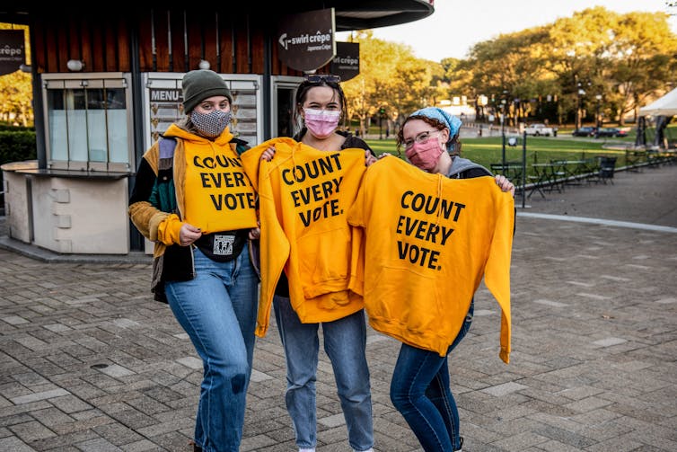 Three young women wear face masks and hold yellow sweatshirts up to them that say 'count every vote'
