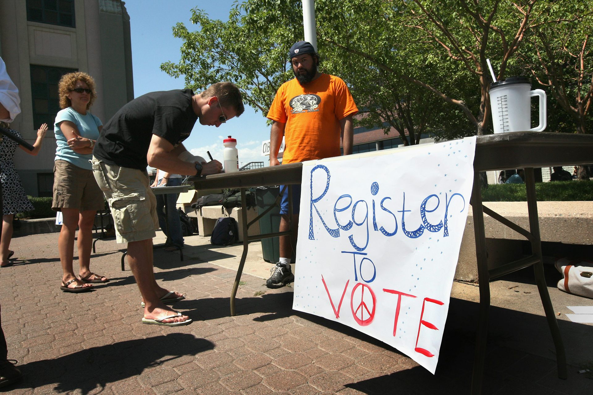 A man in an orange shirt stands next to a table outdoors with a sign that says 'register to vote' while another man leans over the table, filing out paperwork.