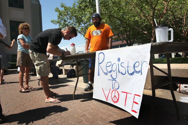 A man in an orange shirt stands next to a table outdoors with a sign that says 'register to vote' while another man leans over the table, filing out paperwork.