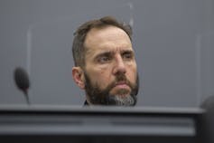 A man with dark hair and a salt-and-pepper beard, sitting behind a large table or desk.