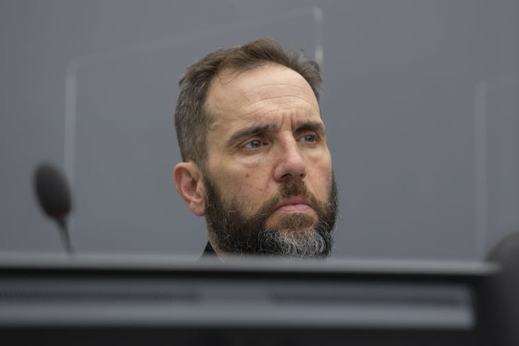 A man with dark hair and a salt-and-pepper beard, sitting behind a large table or desk.