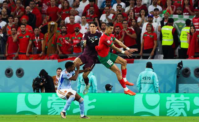 A goalkeeper in purple and defender in red and green jump in the air while a player in blue cowers below them.