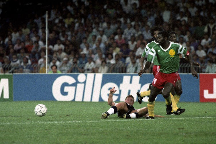 A soocer player is on the ground while another in green jersey looks on.