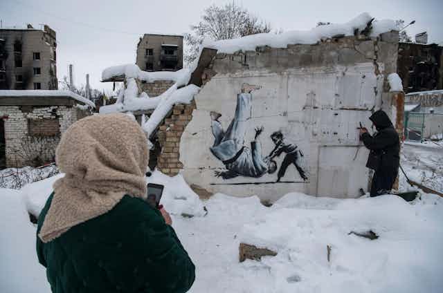A Ukrainian woman watches her friend take a picture of a graffiti by Banksy on a damaged wall in Borodianka, Ukraine, on December 6, 2022