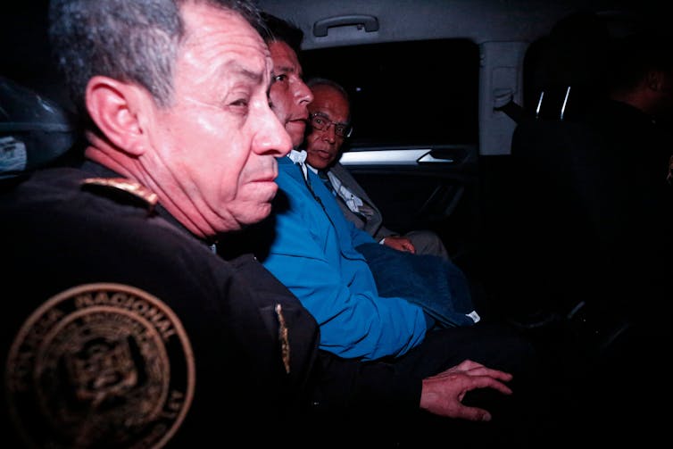 A man in blue is seen inside a car with a police officer next to him in uniform