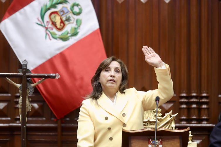 A women in a yellow jacket raises her right hand in front of a Peruvian flag.