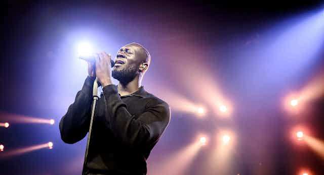 Stormzy dressed all in black on a backlit stage sings into a microphone held on a mic stand.