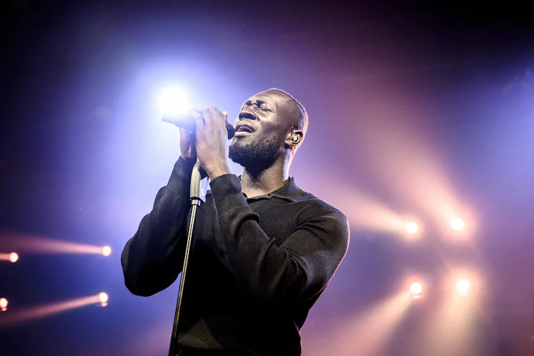 Stormzy dressed all in black on a backlit stage sings into a microphone held on a mic stand.