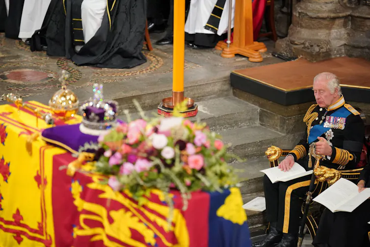 A coffin draped in the royal standard with a purple cushion, a candle and a crown, and the king seated beside it.