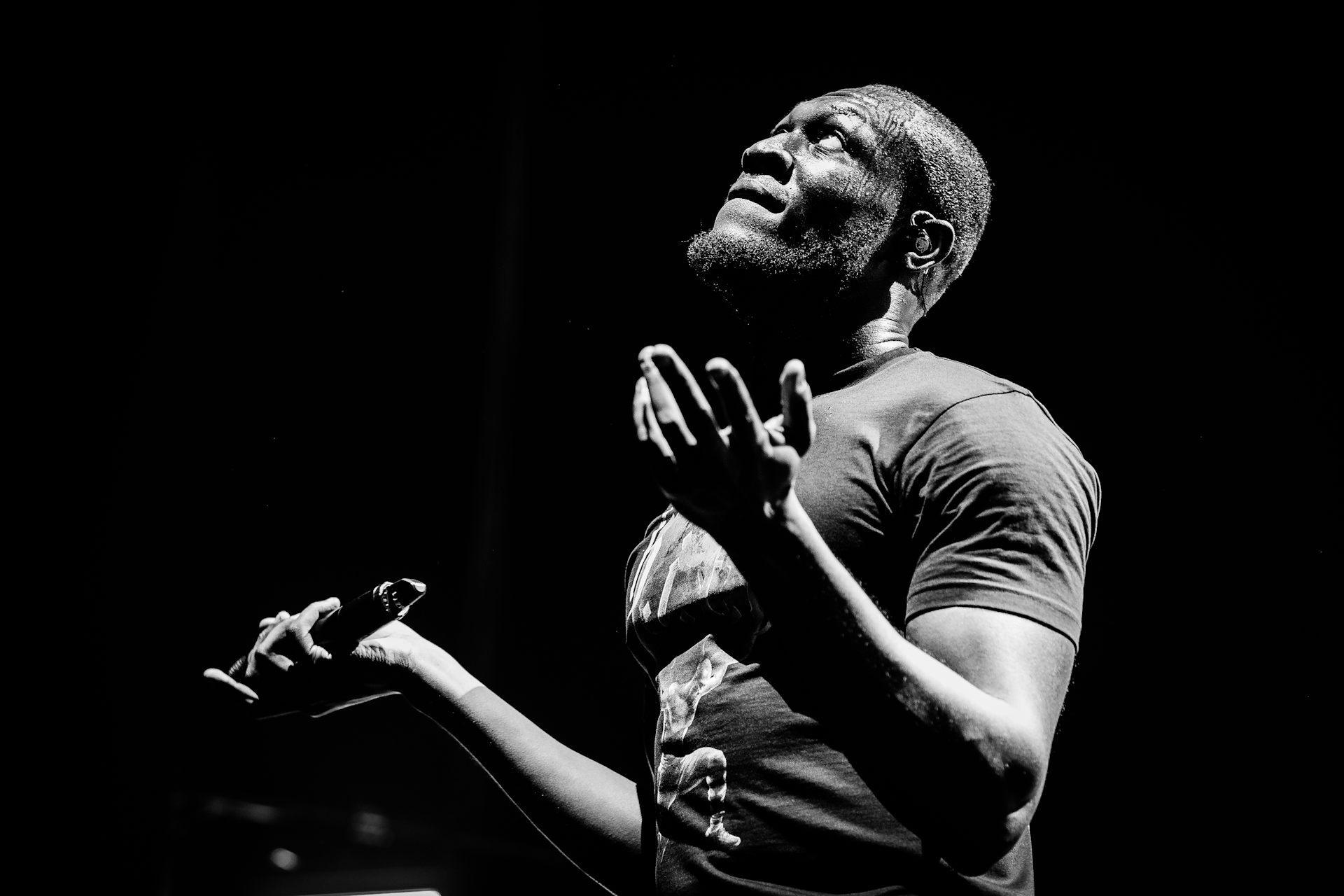 Black and white photo of Stormzy in a t-shirt looking to the sky, his hands outstretched, a microphone in his right.