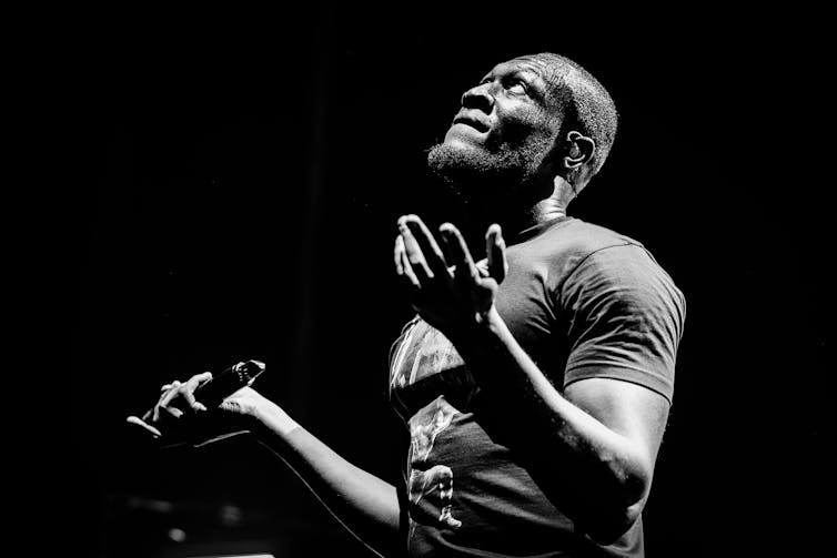 Black and white photo of Stormzy in a t-shirt looking to the sky, his hands outstretched, a microphone in his right.